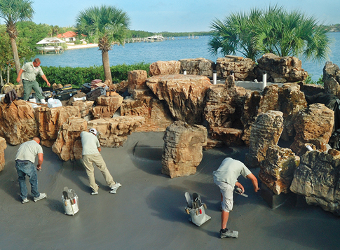 A black pebble plaster finish is being installed in front of the partially completed waterfall structure. 
